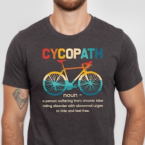 May include: A dark heather grey t-shirt with a vintage-style graphic of a bicycle and the text "CYCOPATH noun - a person suffering from chronic bike riding disorder with abnormal urges to ride and feel free."