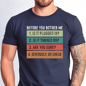 May include: A navy blue t-shirt with a humorous message in a retro style font: "BEFORE YOU BOTHER ME 1. IS IT PLUGGED IN? 2. IS IT TURNED ON? 3. ARE YOU SURE? 4. SERIOUSLY, GO CHECK"