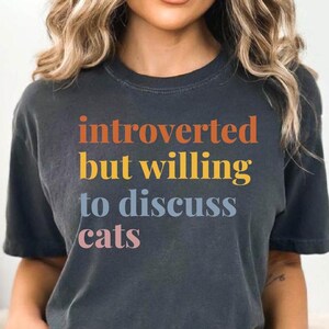 May include: A woman wearing a dark gray t-shirt with the text "introverted but willing to discuss cats" in colorful letters. She is wearing blue denim shorts.