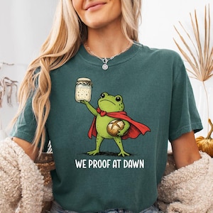 May include: Teal t-shirt featuring a cartoon frog superhero holding a jar and a loaf of bread. The frog wears a red cape and a green bow tie. The text "WE PROOF AT DAWN" is printed below the frog.