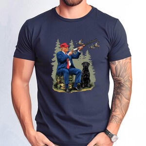 May include: A navy blue t-shirt with a graphic print of a man in a suit and red hat holding a shotgun, with a black dog beside him in a wooded setting.