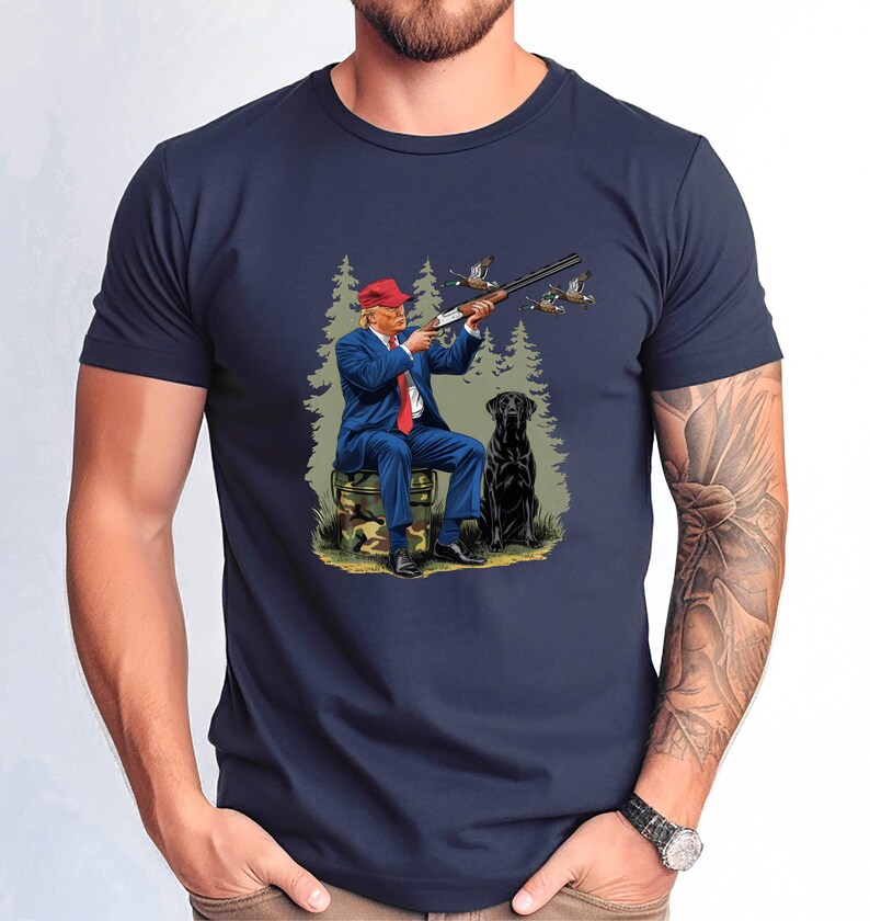 May include: A navy blue t-shirt with a graphic print of a man in a suit and red hat holding a shotgun, with a black dog beside him in a wooded setting.