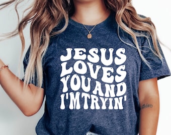 May include: A navy blue t-shirt with white text that reads "JESUS LOVES YOU AND I'M TRYIN'". The text is in a retro, bubble-like font.