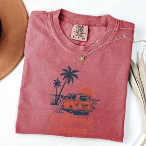 May include: A red t-shirt with a graphic of a vintage orange van, palm trees, and a surfboard. The text "California Dreamin" is printed below the graphic. A gold necklace is draped across the shirt.