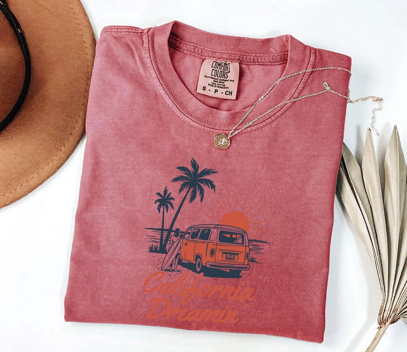 May include: A red t-shirt with a graphic of a vintage orange van, palm trees, and a surfboard. The text "California Dreamin" is printed below the graphic. A gold necklace is draped across the shirt.