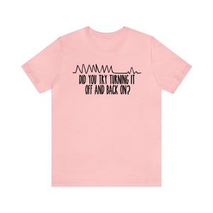 May include: A light pink t-shirt with black text and a graphic of a heartbeat. The text reads "DID YOU TRY TURNING IT OFF AND BACK ON?" The shirt is made of a soft material and has short sleeves.