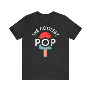 Puede incluir: Camiseta gris oscuro con el texto "THE COOLEST POP" sobre un gr&aacute;fico de paleta roja y azul. La paleta tiene un palo marr&oacute;n. La camiseta est&aacute; hecha de un material suave.
