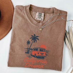 May include: A brown Comfort Colors t-shirt with a graphic of a red van, palm trees, and a setting sun. The text "California Dreamin'" is printed in red. A silver necklace with a pendant is draped on the shirt.