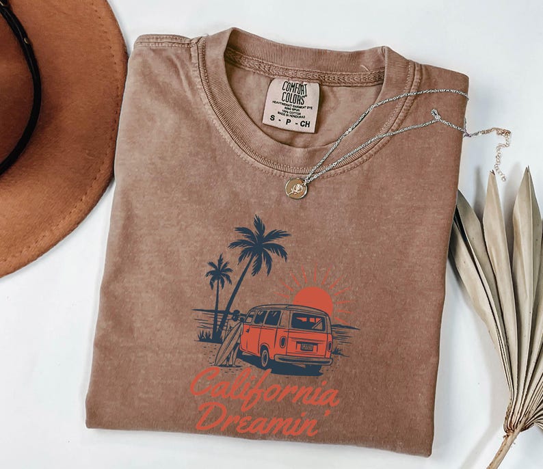 May include: A brown Comfort Colors t-shirt with a graphic of a red van, palm trees, and a setting sun. The text "California Dreamin'" is printed in red. A silver necklace with a pendant is draped on the shirt.