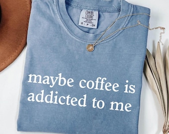 May include: A blue Comfort Colors t-shirt with the phrase "maybe coffee is addicted to me" in white. A gold necklace with a pendant is draped across the shirt. A brown hat and dried plant stems are in the background.