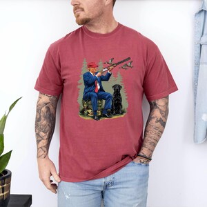 May include: A red t-shirt with a graphic print of a man in a red hat holding a shotgun and a black dog. The man is wearing a blue suit and is sitting on a log. There are ducks flying in the background.