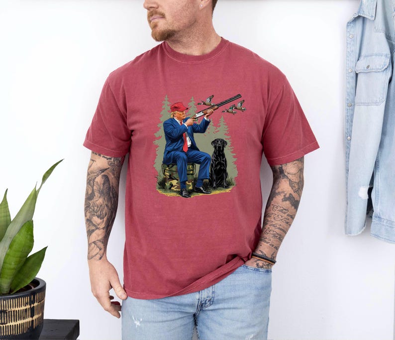 May include: A red t-shirt with a graphic print of a man in a red hat holding a shotgun and a black dog. The man is wearing a blue suit and is sitting on a log. There are ducks flying in the background.