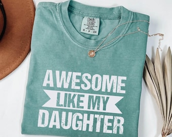 May include: A teal blue t-shirt with the text "Awesome Like My Daughter" in white distressed lettering.