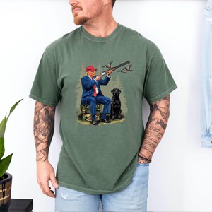 May include: A green t-shirt with a graphic print of a man in a red hat and blue suit holding a shotgun and a black dog. The man is sitting on a log and there are ducks flying in the background.