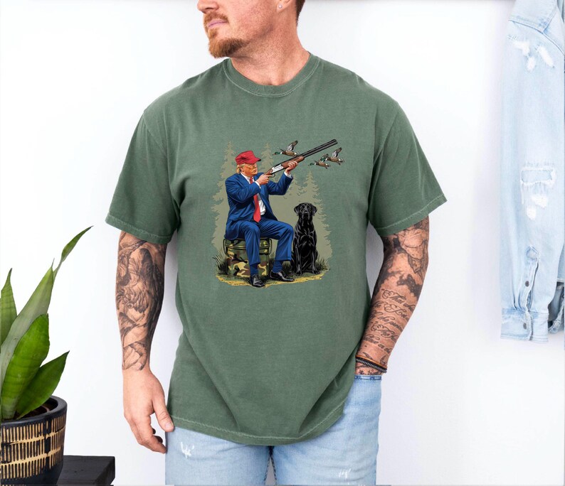 May include: A green t-shirt with a graphic print of a man in a red hat and blue suit holding a shotgun and a black dog. The man is sitting on a log and there are ducks flying in the background.