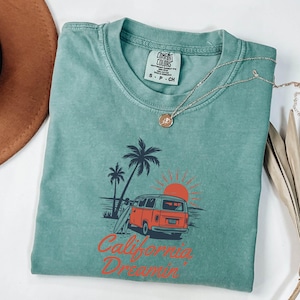 May include: A teal Comfort Colors t-shirt with a graphic of a red and orange van, palm trees, and a setting sun. The text "California Dreamin'" is printed in red. A gold necklace with a pendant is draped on the shirt.