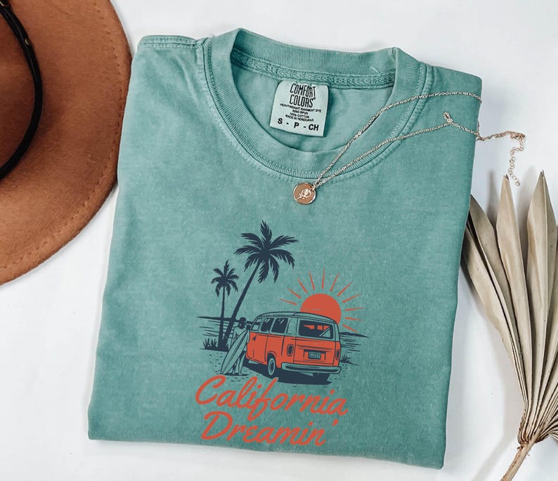May include: A teal Comfort Colors t-shirt with a graphic of a red and orange van, palm trees, and a setting sun. The text "California Dreamin'" is printed in red. A gold necklace with a pendant is draped on the shirt.