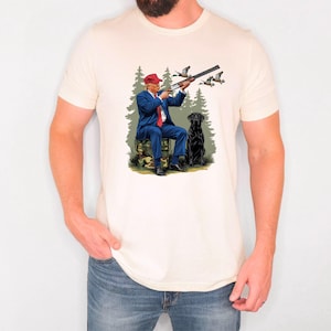 May include: A white t-shirt with a graphic print of a man in a blue suit and red hat holding a shotgun, with a black dog sitting beside him in a forest setting.