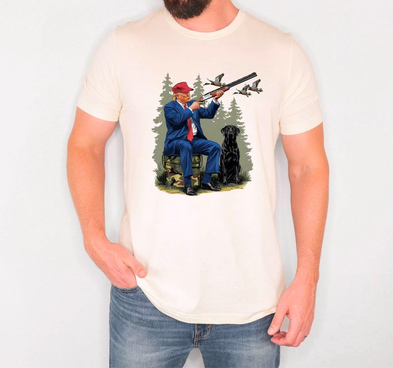 May include: A white t-shirt with a graphic print of a man in a blue suit and red hat holding a shotgun, with a black dog sitting beside him in a forest setting.