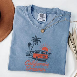 May include: A blue Comfort Colors t-shirt with a graphic of a vintage orange van, palm trees, and a setting sun. The text "California Dreamin'" is printed in orange. A gold necklace with a pendant is draped on the shirt.