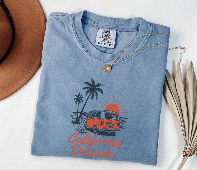 May include: A blue Comfort Colors t-shirt with a graphic of a vintage orange van, palm trees, and a setting sun. The text "California Dreamin'" is printed in orange. A gold necklace with a pendant is draped on the shirt.