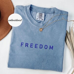 May include: A pale blue t-shirt with the word "FREEDOM" embroidered in blue. The shirt has a crew neck and a small tag on the collar. A gold necklace with a pendant is draped across the shirt.