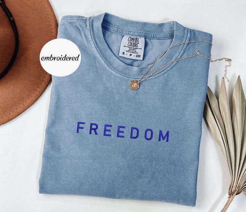 May include: A pale blue t-shirt with the word "FREEDOM" embroidered in blue. The shirt has a crew neck and a small tag on the collar. A gold necklace with a pendant is draped across the shirt.