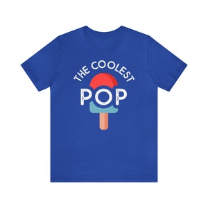 Puede incluir: Camiseta azul real con el texto "THE COOLEST POP" sobre un gr&aacute;fico de paleta. La paleta tiene una parte superior roja, un medio azul claro y un palo marr&oacute;n. El texto y el gr&aacute;fico son blancos.