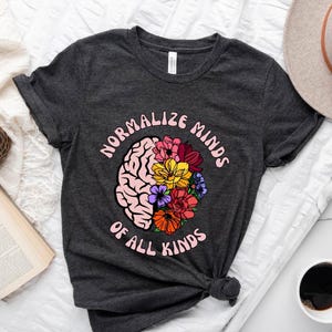 May include: Dark gray t-shirt with the text "NORMALIZE MINDS OF ALL KINDS" in pink. The design features a pink brain graphic with colorful flowers. The shirt is made of a soft material and has short sleeves.