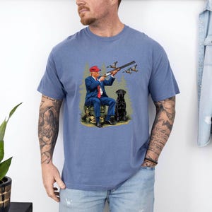 May include: A blue t-shirt with a graphic of a man in a red hat holding a shotgun and a black dog. The man is wearing a blue suit and a red tie. The dog is sitting next to him. The graphic is surrounded by a green forest background.