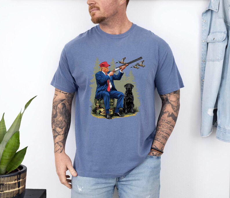 May include: A blue t-shirt with a graphic of a man in a red hat holding a shotgun and a black dog. The man is wearing a blue suit and a red tie. The dog is sitting next to him. The graphic is surrounded by a green forest background.