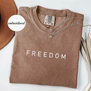 May include: A brown Comfort Colors t-shirt with the word "FREEDOM" printed in white. A silver necklace with a pendant is draped across the shirt. A brown hat and dried plant are in the background. The shirt is size small.