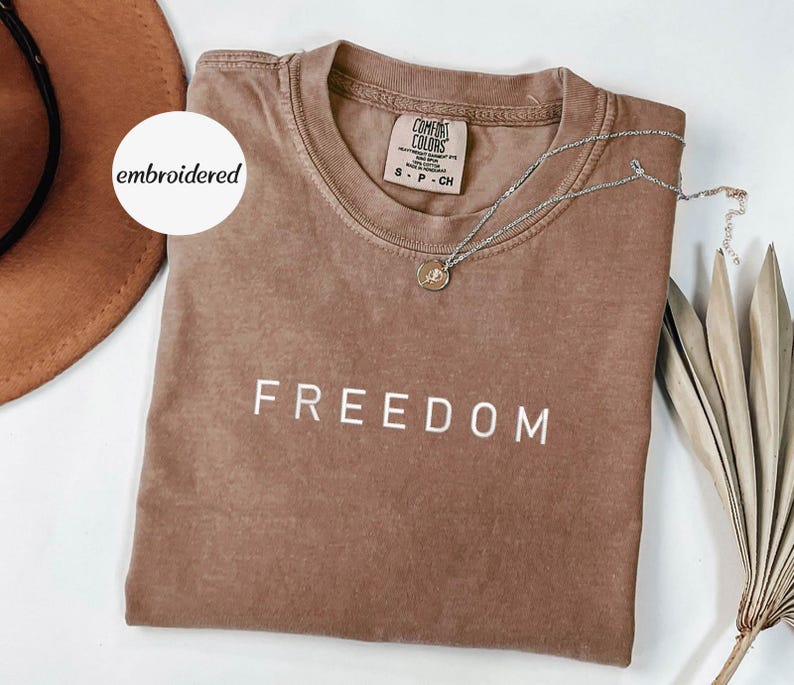 May include: A brown Comfort Colors t-shirt with the word "FREEDOM" printed in white. A silver necklace with a pendant is draped across the shirt. A brown hat and dried plant are in the background. The shirt is size small.