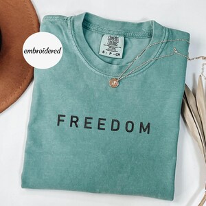 May include: A teal Comfort Colors t-shirt with the word "FREEDOM" printed in black. A gold necklace with a pendant rests on the shirt. The shirt is folded, and a brown hat is visible in the background. The word "embroidered" is on a white circle.