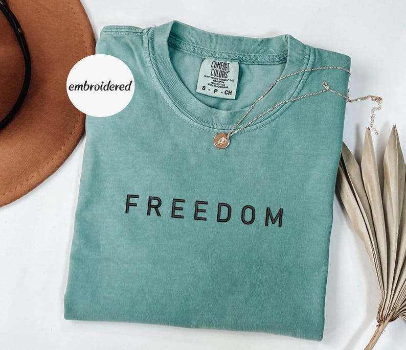 May include: A teal Comfort Colors t-shirt with the word "FREEDOM" printed in black. A gold necklace with a pendant rests on the shirt. The shirt is folded, and a brown hat is visible in the background. The word "embroidered" is on a white circle.