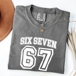 May include: A gray t-shirt with the words "SIX SEVEN" and the number "67" in white block letters. A silver necklace with a pendant is on the shirt. The shirt is folded and next to a brown hat and dried plant.