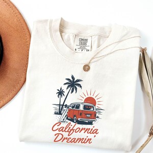 May include: Cream-colored t-shirt featuring a graphic of a red and white van on a beach with palm trees and a setting sun. The text "California Dreamin'" is printed in red script below the image. A gold necklace is draped on the shirt.