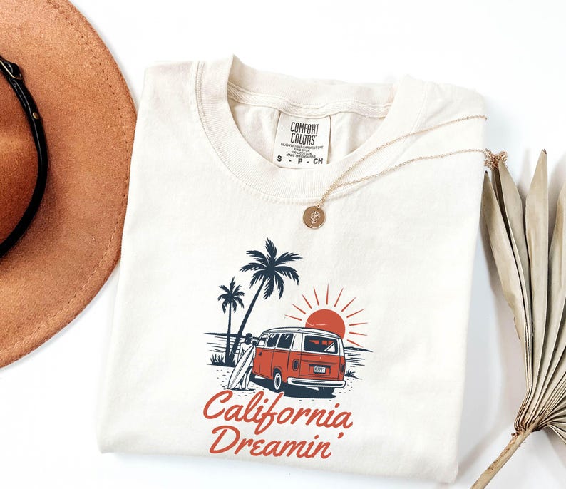 May include: Cream-colored t-shirt featuring a graphic of a red and white van on a beach with palm trees and a setting sun. The text "California Dreamin'" is printed in red script below the image. A gold necklace is draped on the shirt.