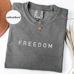 May include: A grey Comfort Colors t-shirt with the word "FREEDOM" in white block letters. A gold necklace with a pendant rests on the shirt. The shirt is folded and displayed with a brown hat and dried plant.