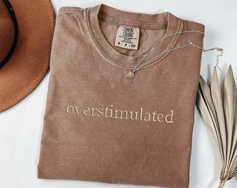 May include: A brown t-shirt with the text "overstimulated" embroidered in white on the front. The shirt is made of a soft, comfortable material and has a relaxed fit.