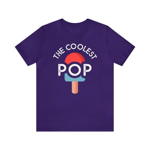 Puede incluir: Una camiseta morada con las palabras "THE COOLEST POP" en blanco sobre un gr&aacute;fico de paleta. La paleta tiene una parte superior roja, un centro azul y un palo marr&oacute;n. La camiseta est&aacute; hecha de un material suave.