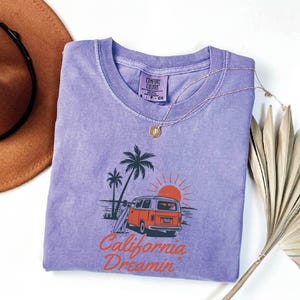 May include: A lavender Comfort Colors t-shirt featuring a graphic of a vintage orange van, palm trees, and a setting sun. The text "California Dreamin" is printed in orange. A gold necklace and a brown hat are also visible.