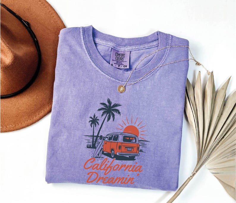 May include: A lavender Comfort Colors t-shirt featuring a graphic of a vintage orange van, palm trees, and a setting sun. The text "California Dreamin" is printed in orange. A gold necklace and a brown hat are also visible.