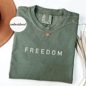 May include: An olive green t-shirt with the word "FREEDOM" embroidered in white. A gold necklace with a pendant rests on the shirt. The shirt is folded, and a brown hat is visible in the background.