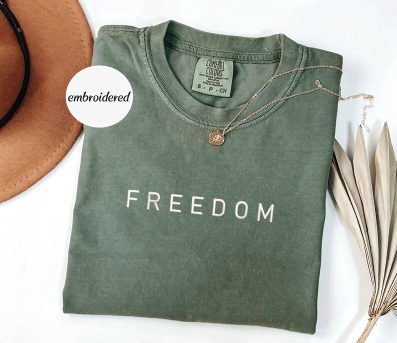 May include: An olive green t-shirt with the word "FREEDOM" embroidered in white. A gold necklace with a pendant rests on the shirt. The shirt is folded, and a brown hat is visible in the background.