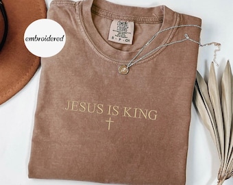 May include: A brown Comfort Colors t-shirt with the embroidered text "JESUS IS KING" and a cross. A silver necklace with a pendant rests on the shirt. The shirt is folded and placed on a white surface.