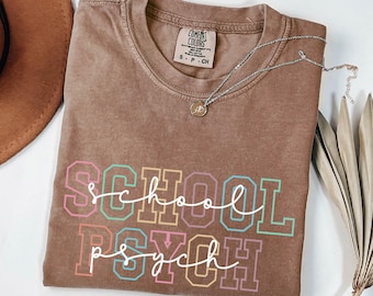 May include: A brown Comfort Colors t-shirt with the words "SCHOOL PSYCH" in a colorful, collegiate-style font. A silver necklace with a small pendant is draped across the shirt. A brown hat and dried leaves are in the background.