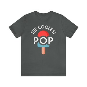 Puede incluir: Camiseta gris oscuro con el texto "THE COOLEST POP" sobre un gr&aacute;fico de paleta de helado roja, azul y naranja. La camiseta es de cuello redondo y manga corta.