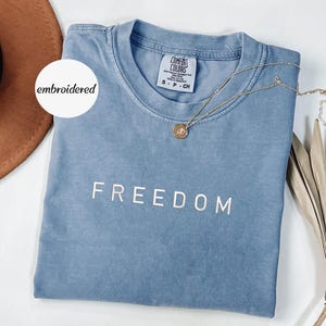 May include: A blue t-shirt with the word "FREEDOM" printed in white. A gold necklace with a pendant rests on the shirt. The shirt has a tag that reads "COMFORT COLORS". A white circle with the word "embroidered" is in the upper left corner.