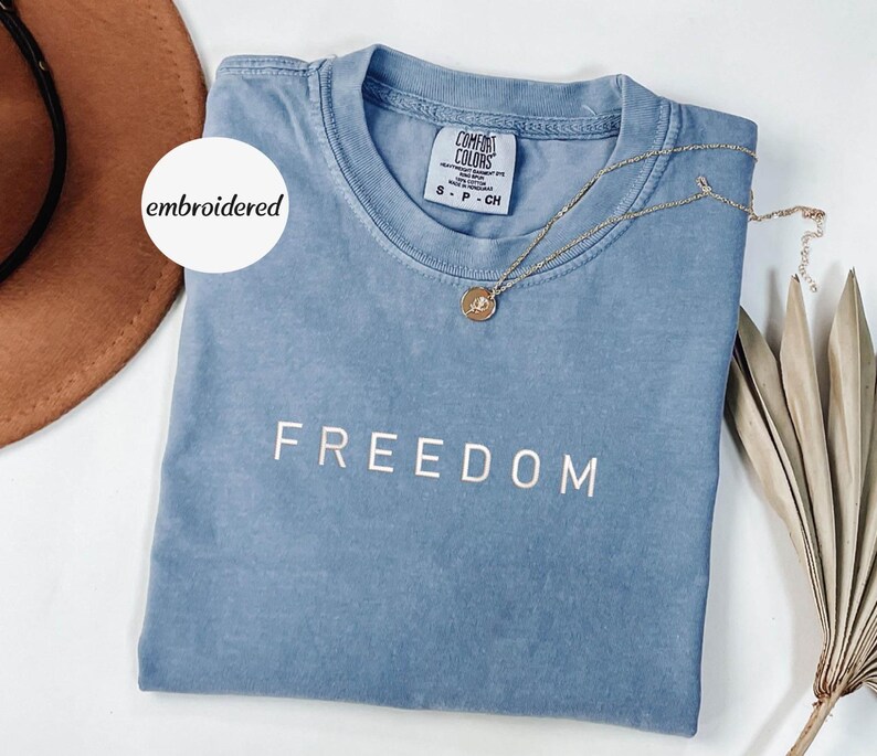 May include: A blue t-shirt with the word "FREEDOM" printed in white. A gold necklace with a pendant rests on the shirt. The shirt has a tag that reads "COMFORT COLORS". A white circle with the word "embroidered" is in the upper left corner.